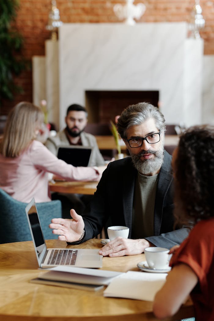 A Man in Black Suit Wearing Eyeglasses Sitting Near the Table while Talking to His Colleague